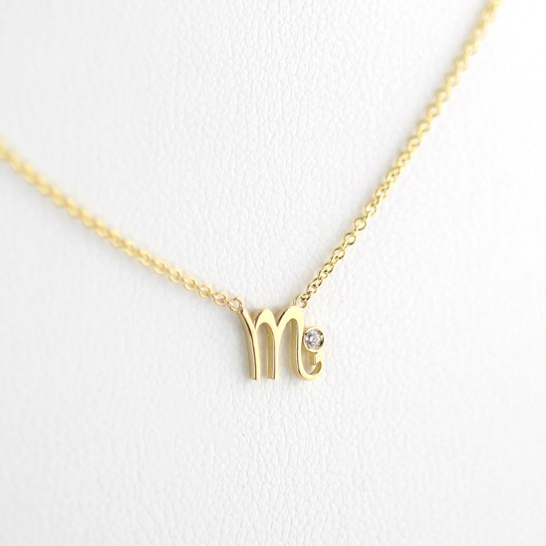 Puede incluir: Un collar de oro con un peque&ntilde;o acento de diamante y un colgante en forma de la letra "M".