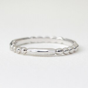 May include: A silver ring with a unique design. The ring features a series of small, round beads along the band, with a section of small diamonds and a flat, rectangular center. The ring is set against a white background.