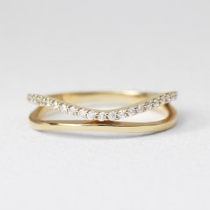 May include: A gold-colored ring with a double-band design. The top band curves and is adorned with small, clear gemstones. The lower band is a smooth, solid gold. The ring is set against a white background, highlighting its elegant simplicity.