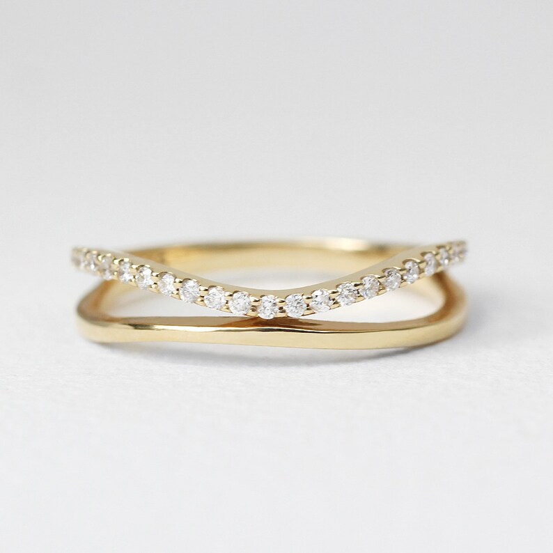 May include: A gold-colored ring with a double-band design. The top band curves and is adorned with small, clear gemstones. The lower band is a smooth, solid gold. The ring is set against a white background, highlighting its elegant simplicity.