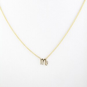 Puede incluir: Un collar de cadena de oro con un peque&ntilde;o colgante en forma de la letra "M" con un solo acento de diamante.