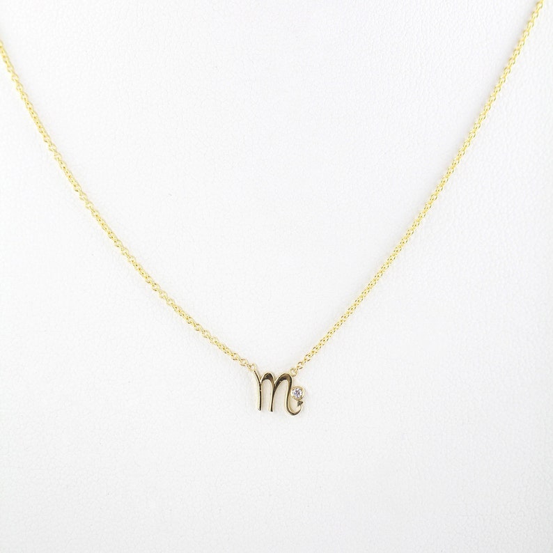 Puede incluir: Un collar de cadena de oro con un peque&ntilde;o colgante en forma de la letra "M" con un solo acento de diamante.