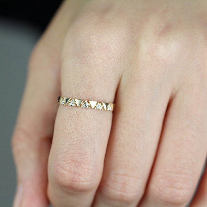 Pu&ograve; includere: Un anello color oro con un motivo ripetuto di triangoli e piccole gemme trasparenti. L'anello &egrave; indossato su un dito, mostrando il suo design delicato e la sua maestria artigianale. La fascia dell'anello &egrave; sottile ed elegante.