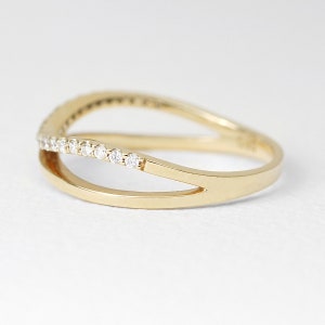 May include: A gold ring with a split band design. The top band is adorned with a row of small, clear gemstones. The ring has a polished finish and a modern, elegant style. The ring is set against a white background.