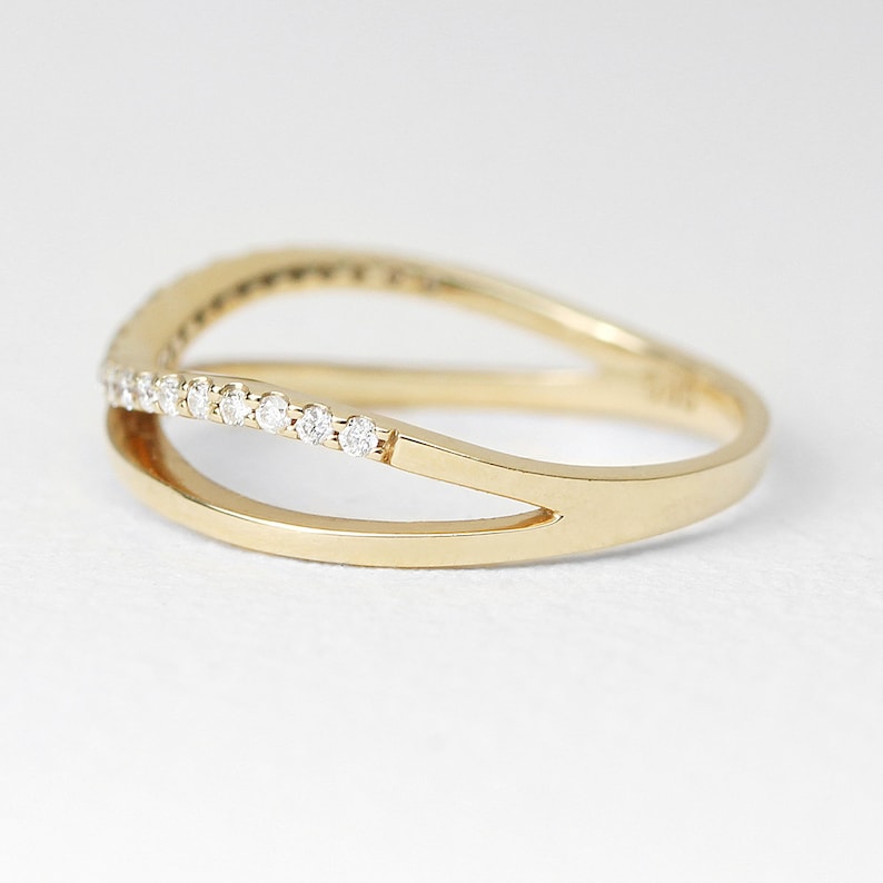 May include: A gold ring with a split band design. The top band is adorned with a row of small, clear gemstones. The ring has a polished finish and a modern, elegant style. The ring is set against a white background.