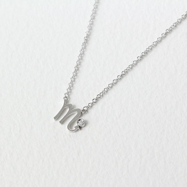 Puede incluir: Un collar de plata con una peque&ntilde;a piedra preciosa transparente y un colgante con la letra 'M' estilizada.