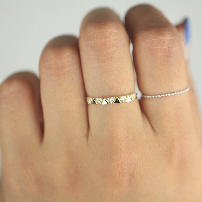 Pu&ograve; includere: Un anello color oro con un motivo ripetuto di piccoli triangoli, alcuni riempiti di smalto bianco e nero, e altri con piccole pietre trasparenti. &Egrave; visibile anche un secondo anello sottile con piccole pietre trasparenti.