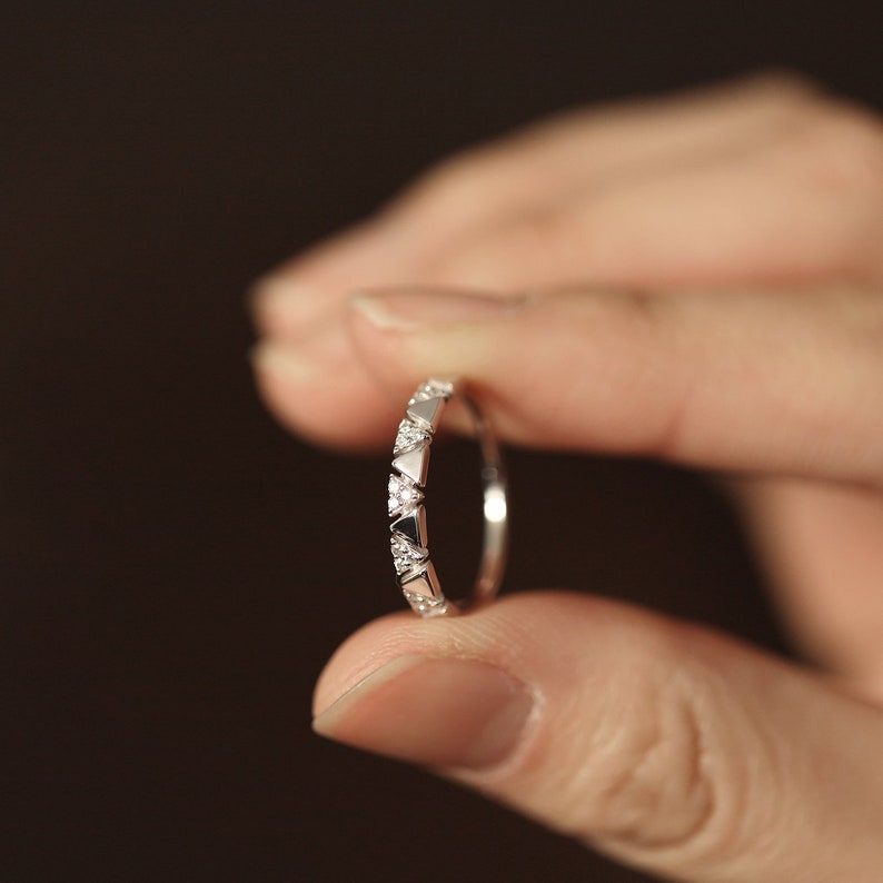 Pu&ograve; includere: Un delicato anello d'argento con un motivo ripetuto di accenti triangolari e a forma di diamante. L'anello &egrave; tenuto tra due dita su uno sfondo sfocato, evidenziando il suo design intricato e l'elegante semplicit&agrave;.