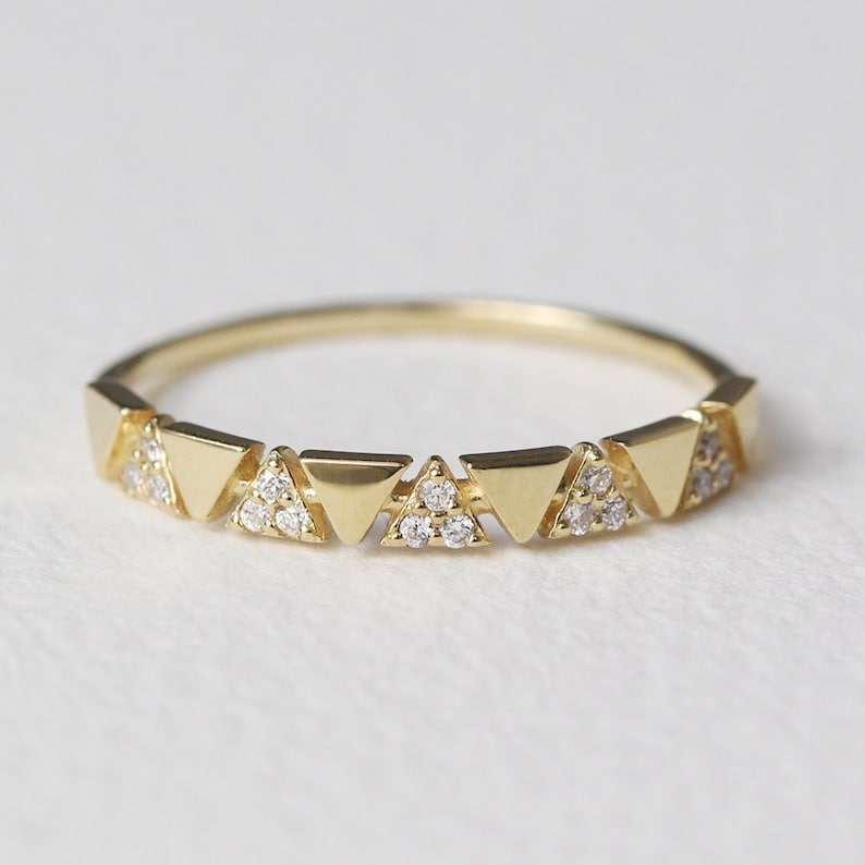 Pu&ograve; includere: Un anello dorato con una serie di forme triangolari. Alcuni triangoli sono adornati con piccole gemme scintillanti. L'anello ha una fascia delicata e un design geometrico moderno.