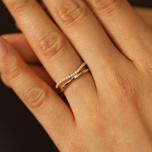 May include: A gold-colored ring with a crisscross design. One band is adorned with small, clear gemstones. The ring is on a finger, with a neutral background.