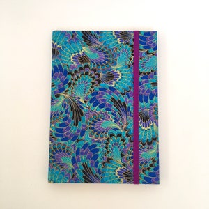 May include: A rectangular journal with a vibrant, patterned cover. The design features swirling peacock feather-like shapes in shades of blue, purple, and gold against a black background. A purple elastic band secures the journal.