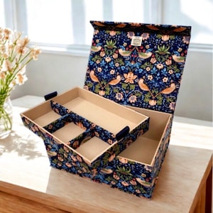 May include: A rectangular storage box with a hinged lid, covered in a dark blue fabric featuring a repeating pattern of orange birds, green foliage, and light pink flowers. The interior has a removable beige tray with four compartments.