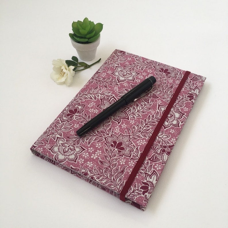 May include: A journal with a floral pattern in shades of pink and white, featuring a matching elastic band and a black pen. The journal is closed, with the pen resting on top. A small potted succulent and a white flower are in the background.