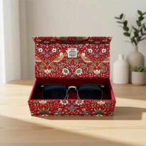May include: A red fabric-covered storage box with a hinged lid featuring a repeating pattern of birds and flowers. The interior is lined with red velvet and contains a pair of black-rimmed sunglasses. A small white label reads Upcrafts London.