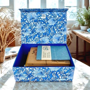 Peut inclure: Coffre de rangement bleu orné d'un motif floral bleu et blanc. À l'intérieur, plusieurs livres et carnets. Le coffre est posé sur une table près d'une fenêtre.