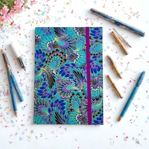 May include: A journal with a teal, blue, purple, and gold feather pattern and a magenta elastic closure. Several pens in various styles and colors are scattered around the journal, including blue and gold pens and bamboo-style pens.