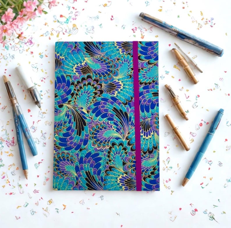 May include: A journal with a teal, blue, purple, and gold feather pattern and a magenta elastic closure. Several pens in various styles and colors are scattered around the journal, including blue and gold pens and bamboo-style pens.
