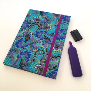 May include: A closed notebook with a vibrant blue, teal, and purple floral pattern, gold accents, and a purple elastic band. A purple highlighter with a black cap is next to the notebook. Stationery.