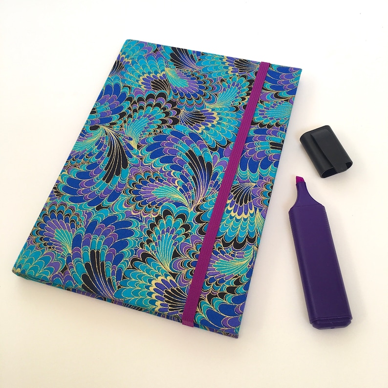 May include: A closed notebook with a vibrant blue, teal, and purple floral pattern, gold accents, and a purple elastic band. A purple highlighter with a black cap is next to the notebook. Stationery.
