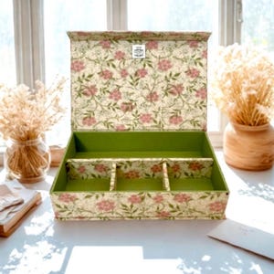 Puede incluir: Una caja de almacenamiento rectangular con tapa abatible, que presenta un exterior de color crema con un patrón de flores rosas y hojas verdes. El interior está forrado en verde sólido e incluye tres divisores con el mismo patrón floral.