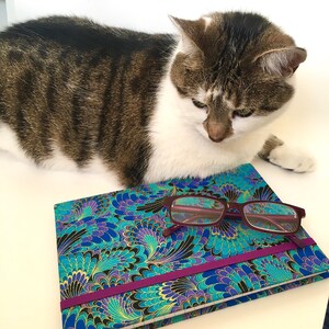 May include: A patterned journal with a teal, blue, and gold design, a purple elastic band, and a pair of maroon eyeglasses. A tabby cat is lying above the journal. The journal's cover features a floral pattern.
