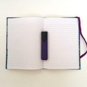 May include: An open notebook with lined pages and a purple highlighter. The notebook has a patterned cover with blue, green, and gold accents. A purple ribbon bookmark is attached to the spine.