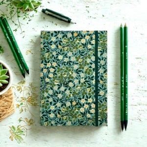 May include: A notebook with a teal floral design, green elastic closure, and pencils. The cover features a pattern of white and yellow flowers with green foliage. A small succulent plant in a white pot is also visible.