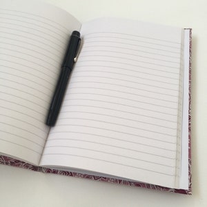 May include: An open notebook with lined pages and a black pen resting on the left side. The notebook has a patterned border in shades of purple and white. The pen is black with a silver clip.
