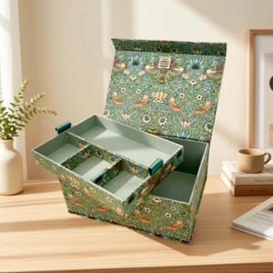 Può includere: Una scatola rettangolare con coperchio incernierato, rivestita in tessuto verde con un motivo ripetuto di uccelli e fiori. L'interno è foderato in verde chiaro e include un vassoio rimovibile con tre scomparti separati.