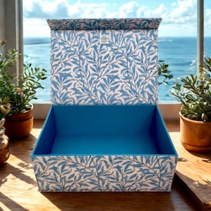 Puede incluir: Una caja rectangular abierta con interior azul y tapa. El exterior presenta un fondo blanco con un patrón de hojas azules. La caja está sobre una superficie de madera, con plantas en macetas y vista al mar al fondo.