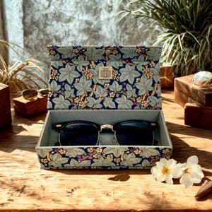 May include: Sunglasses in a decorative case with a floral pattern. The case is open, revealing black sunglasses with gold-coloured rims. The case has a navy blue background with green leaves and small yellow and pink flowers. The case has the text "MORRIS & Co LONDON" on the top.
