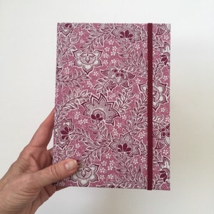 May include: A rectangular journal with a floral pattern in shades of pink and white. The cover features a repeating design of stylized flowers and leaves. A burgundy elastic band secures the journal. The journal is held by a hand.