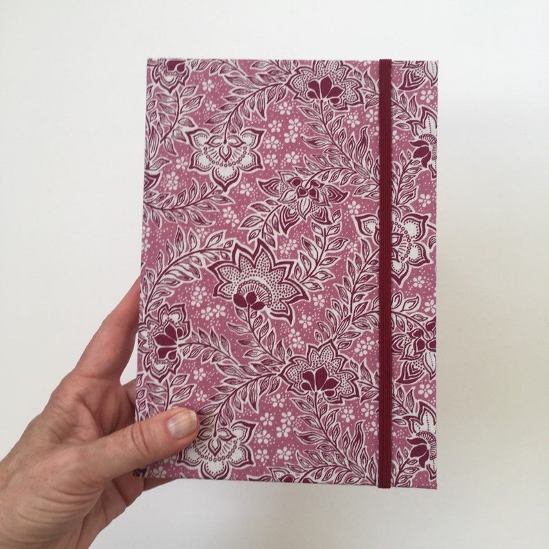 May include: A rectangular journal with a floral pattern in shades of pink and white. The cover features a repeating design of stylized flowers and leaves. A burgundy elastic band secures the journal. The journal is held by a hand.