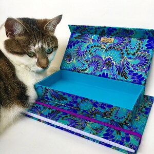 May include: An open, rectangular box and a matching notebook with a blue, black, and gold floral pattern. The box is turquoise inside. The notebook has a purple elastic band. The box has the text "UNICRAFTS". A cat is next to the box.
