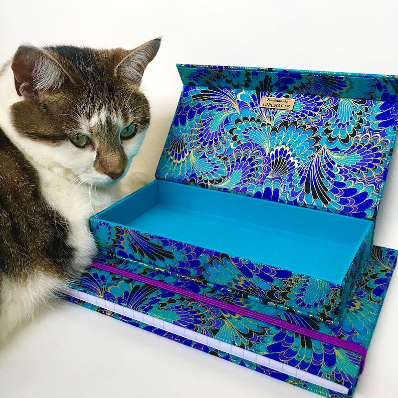May include: An open, rectangular box and a matching notebook with a blue, black, and gold floral pattern. The box is turquoise inside. The notebook has a purple elastic band. The box has the text "UNICRAFTS". A cat is next to the box.