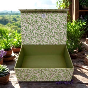 Puede incluir: Una caja decorativa abierta con interior verde y tapa. El exterior presenta un patrón repetido de hojas verdes sobre un fondo rosa claro. La caja está sobre una superficie de madera, con plantas en el fondo.