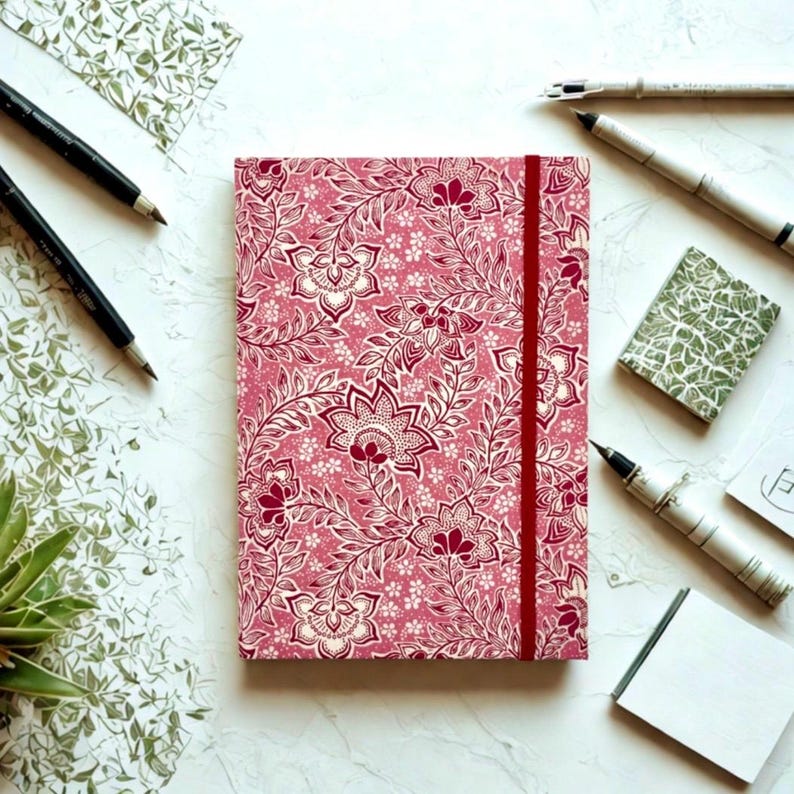 May include: A pink journal with a floral pattern and a red elastic band closure. The journal is surrounded by pens, pencils, and small notepads on a white surface. The floral design features white and burgundy accents.