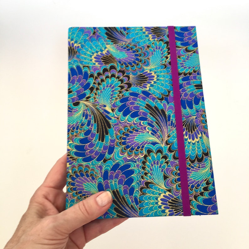 May include: A hardback journal with a vibrant, patterned cover. The design features a peacock feather motif in shades of blue, teal, purple, and gold. A magenta elastic band secures the journal. The journal is held in a hand.