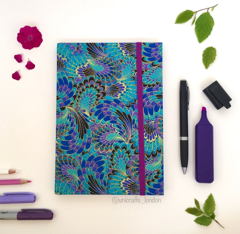 May include: A notebook with a blue, purple, and gold feather pattern cover and a purple elastic band. A black pen, purple highlighter, and other stationery items are arranged around the notebook. The text "@unicrafts_london" is visible.