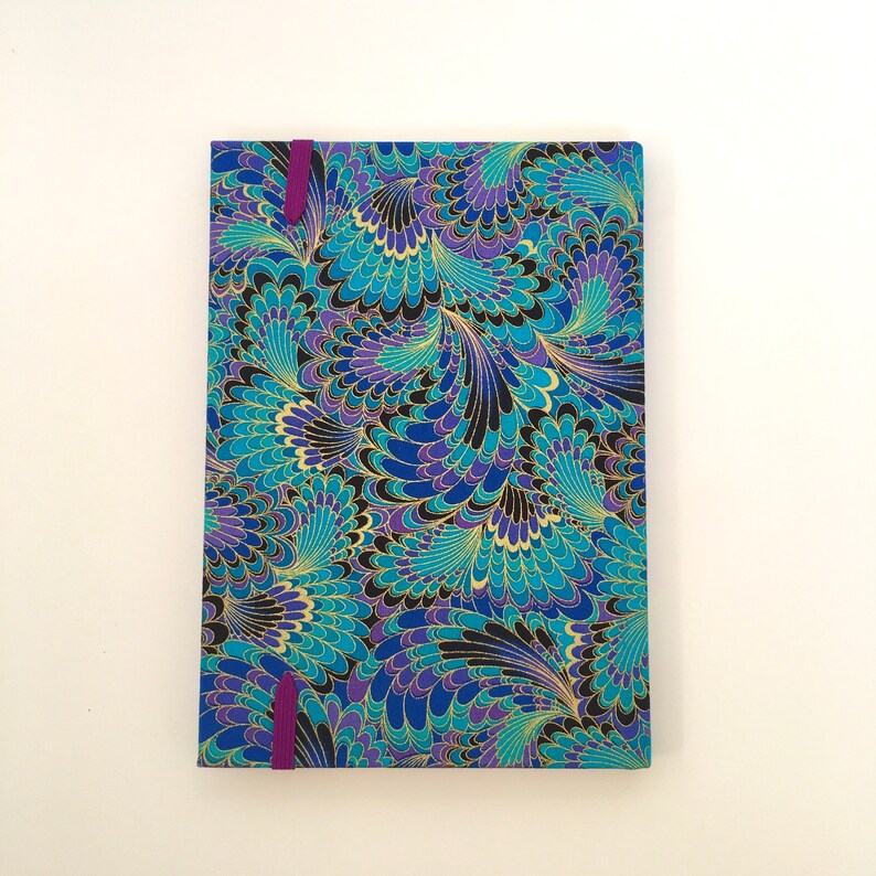 May include: A rectangular journal with a vibrant, patterned cover. The design features swirling peacock feather-like shapes in shades of blue, teal, purple, and gold against a black background. A purple ribbon bookmark is visible. The journal is a stationery item.