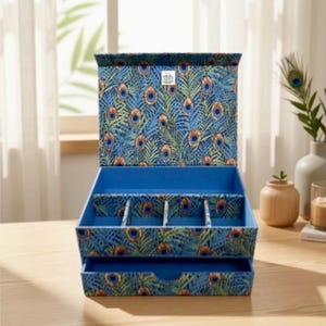 May include: A rectangular jewellery box featuring a vibrant blue and green peacock feather pattern. The box has an open lid, a top compartment divided into four sections, and a single pull-out drawer at the bottom, all lined in solid blue.