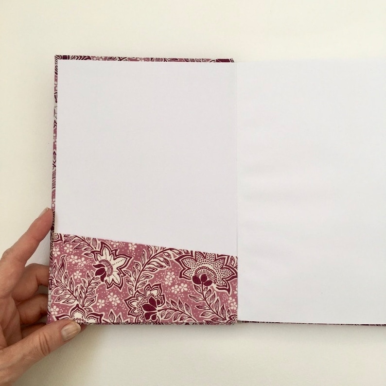 May include: Open notebook with a floral patterned cover and pocket. The cover and pocket feature a design with burgundy and white flowers and leaves on a mauve background. The notebook is open to reveal blank white pages.