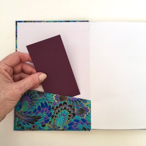May include: A handmade journal with a decorative pocket. The pocket features a blue, teal, and gold floral pattern. A hand is inserting a burgundy card into the pocket. The journal pages are white.