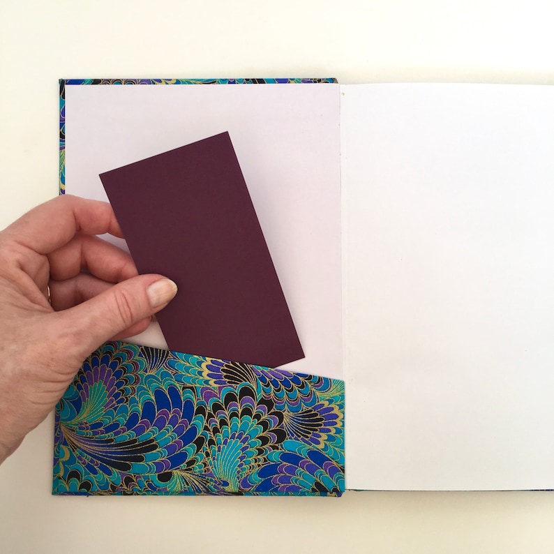 May include: A handmade journal with a decorative pocket. The pocket features a blue, teal, and gold floral pattern. A hand is inserting a burgundy card into the pocket. The journal pages are white.