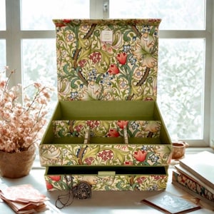 Peut inclure: Une boîte à bijoux ouverte avec un motif floral vert, rouge et bleu sur fond crème. L'intérieur est doublé de tissu vert. La boîte comporte plusieurs compartiments et un tiroir. Un collier avec un pendentif en forme de cœur repose sur la surface.