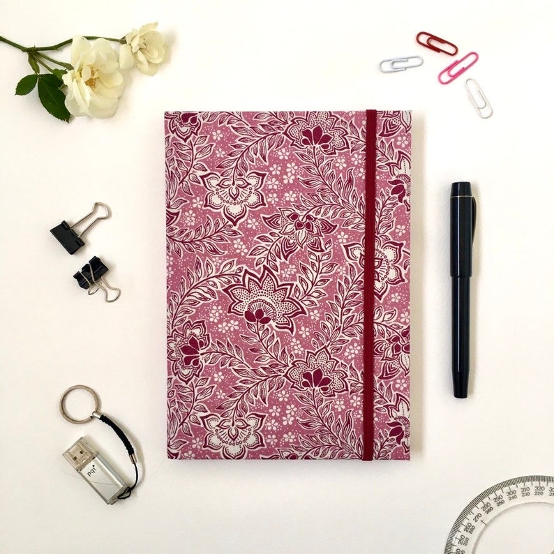 May include: A floral patterned journal with a burgundy elastic band and matching floral design. A black pen, paper clips, and a USB drive keychain are arranged around the journal on a white surface. White flowers are in the upper left corner.