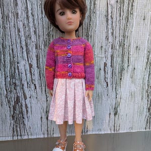 May include: A doll wearing a pink, purple, and orange striped cardigan with purple buttons, a pink floral skirt, and white shoes with brown straps. The doll has brown hair and is standing in front of a weathered, white wooden background.