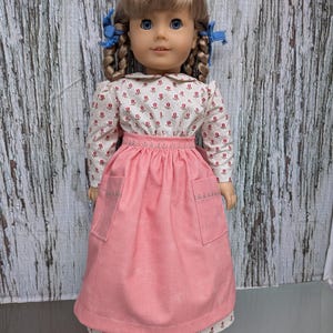 Puede incluir: Muñeca con trenzas rubias y ojos azules, vestida con un vestido rosa. El vestido tiene un corpiño blanco con estampado floral, una falda rosa, un cuello blanco y dos bolsillos. La muñeca lleva zapatos marrones.