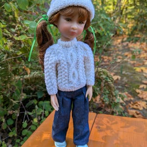 May include: A doll with auburn hair in pigtails tied with green ribbons, wearing a white cable-knit jumper, a matching white knit beanie, dark blue denim trousers, and light blue shoes. The doll has brown eyes and a neutral facial expression.