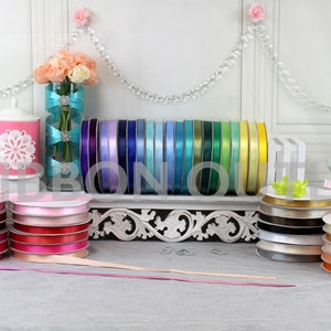 May include: A display of various satin ribbons in a range of colors, including pinks, purples, blues, greens, and yellows. The ribbons are on spools, and some are stacked. A decorative vase with flowers and a pink container are also visible.
